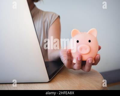 Geldbox zur Hand. Die Hand hält das pinke Sparschwein, während man den Laptop auf dem Schreibtisch benutzt. Geld sparen Reichtum und Finanzkonzept, Geschäft, online b Stockfoto