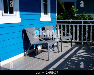 Zwei leere moderne Sessel, graue Farbe mit Beistelltisch auf der Terrasse vor dem Raum des blauen Hauses mit Sonnenlicht. Stockfoto