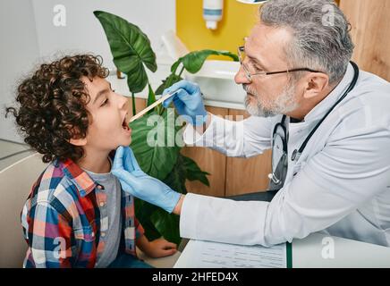 Doktor mit Inspektionsspachtel, um die Kehle eines Jungen aus dem Nahen Osten zu untersuchen. Lockiges männliches Kind beim Kinderarzttermin Stockfoto