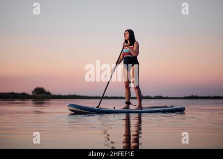 Entspannte Frau mittleren Alters rudert auf dem Brett mit Ruder Blick auf den Sonnenuntergang auf dem See gewelltes Wasser mit rosa Himmel im Hintergrund. Aktiver Lebensstil für ältere Menschen Stockfoto