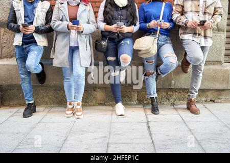Nicht erkennbare männliche und weibliche Freunde in legerer Kleidung, die an der Wand gelehnt ist, und in sozialen Medien auf Mobiltelefonen auf der Straße in der Stadt surfen Stockfoto
