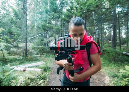 Professioneller Videograf, der eine DSLR-Kamera auf einem 3-Achsen-Gimbal-Stabilisator im Wald hält. Pro Equipment hilft, hochwertige Videos ohne Schütteln zu machen. Kameramann. Zeitlupe Stockfoto