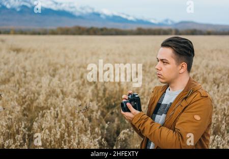 Der Typ ist Fotograf auf dem Feld, vor dem Hintergrund der Berge, macht ein Bild auf einer alten Retro-Filmkamera. Der Mann geht im Herbst in legerer Kleidung. Konzept Lifestyle Millennial Hipster Stockfoto