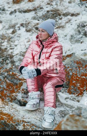 Die Frau sitzt auf dem Hügel. Der Reisende läuft in den oberen Bergen. Schönes Mädchen ist in stilvollen rosa Daunenjacke gekleidet. Explorer schaut weit weg, auf einen schönen Himmel. Magischer reinster Platz in der Natur. Stockfoto