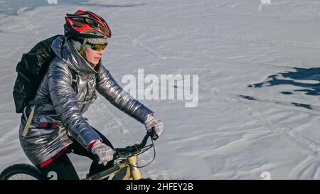 Frau fährt Fahrrad auf dem Eis. Das Mädchen ist in einer silbrigen Daunenjacke, einem Fahrradrucksack und einem Helm gekleidet. Eis des gefrorenen Baikalsees. Reifen auf dem Fahrrad sind mit Spikes bedeckt. Der Reisende fährt mit dem Fahrrad. Stockfoto