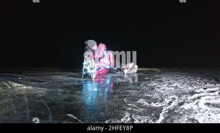 Junge liebende Paar Schlittschuhlaufen auf der Eisbahn in der Nacht. Mann und Frau küssen, umarmen, haben Spaß, lernen zu skaten. Liebe auf Eis machen. Schönes klares Eis mit Rissen am See. Ice leuchtet von unten mit Hintergrundbeleuchtung. Stockfoto