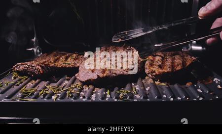 Köstliche saftige Fleisch Steak Kochen auf dem Grill. Küchenzange. Gereifte Prime Rare Braten Grillfilet frischen Marmor Zartheit Rindfleisch. Frittiertes Rindfleisch auf elektrischem Bräter, schwarzer Rosmarin-Pfeffer, Salz. Stockfoto