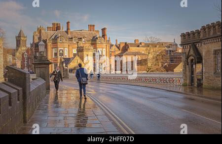 Die Menschen gehen über eine Brücke, die noch nass ist von einem Regenschauer vor kurzem. Alte Gebäude und ein Kirchturm sind auf der anderen Seite und die Leute laufen über das Ufer Stockfoto