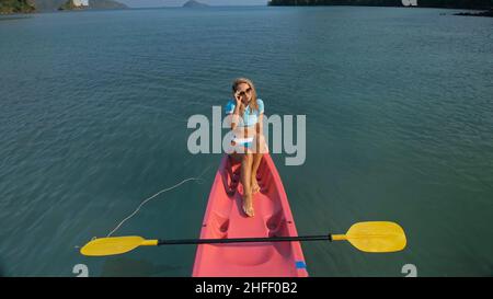 Die ziemlich schlanke Frau im eleganten Badeanzug liegt im rosa Plastikkajak, das auf dem azurblauen Ozean in der exotischen Urlaubsoberansicht driftet. Stockfoto