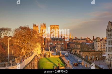 Blick auf das York Minster, das von den historischen Stadtmauern in der Abendsonne gefangen wurde. Alte Gebäude stehen herum und Menschen und Autos überqueren die Brücke. Stockfoto