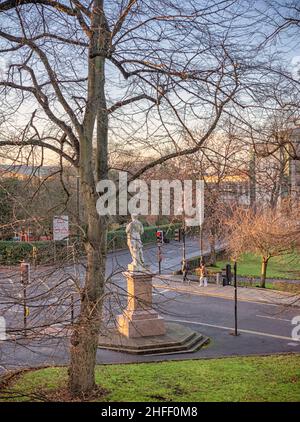 Eine Statue steht an einer Kreuzung mit einem großen Baum im Vordergrund. An der Kreuzung sind Ampeln, und die Leute gehen auf dem Bürgersteig. Stockfoto