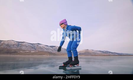 Das Kind trainiert auf Eisschnelllauf. Das Mädchen Schlittschuhe im Winter in Sportbekleidung, Sportbrille, Anzug. Zeitlupe im Freien. Stockfoto