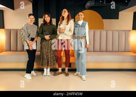 Vier Unternehmerinnen lächeln fröhlich, während sie in einer Bürolobby zusammenstehen. Gruppe von multiethnischen Geschäftsfrauen arbeiten als Team in einem modernen Stockfoto