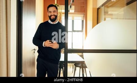 Erfolgreicher junger Geschäftsmann lächelt, während er ein Smartphone in einem modernen Büro hält. Fröhlicher junger Geschäftsmann, der im Stehen eine SMS sendet Stockfoto