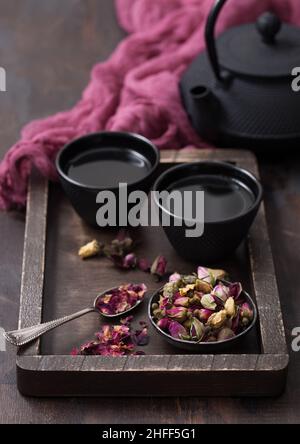 Rose Knospen Tee in Holzkiste mit Löffel und Eisenbecher mit Teekannen und rotem Tuch auf Holz Hintergrund. Stockfoto