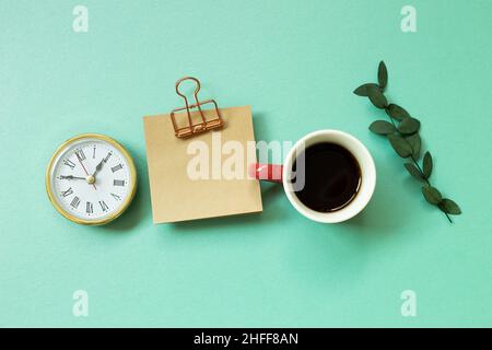 Notizblock, Uhr, Tasse Kaffee, grünes Blatt auf mintgrünem Hintergrund. Schreibtisch, Arbeitsplatz. Draufsicht Stockfoto