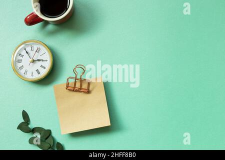 Notizblock, Uhr, Tasse Kaffee, grünes Blatt auf mintgrünem Hintergrund. Schreibtisch, Arbeitsplatz. Draufsicht Stockfoto