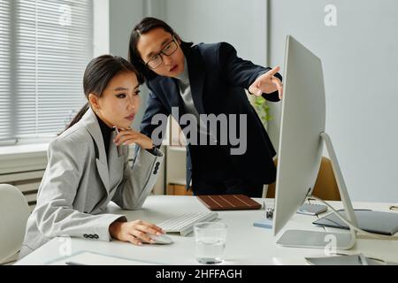Porträt einer jungen asiatischen Geschäftsfrau, die am Schreibtisch im Büro arbeitet, mit einem Manager, der auf den Bildschirm zeigt und Anweisungen gibt und Platz für Kopien gibt Stockfoto