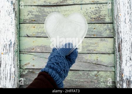 Valentinstag. Herz, ein Symbol der Liebe, aus Eis, wird von einer Hand in Fäustlingen vor dem Hintergrund einer alten Holzwand gehalten. Stockfoto