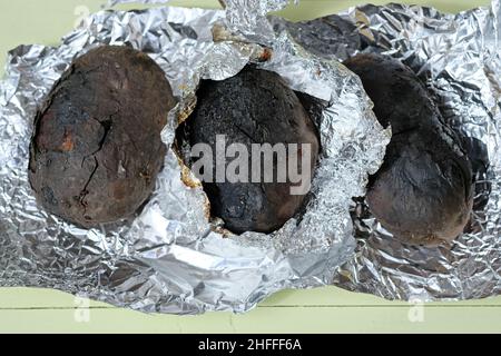 Gebackene Kartoffeln in einer Schale auf Holzkohle. Drei Kartoffeln in Folie über einem Feuer gebacken. Gebackene Kartoffeln mit einer schwarzen, verbrannten Kruste. Stockfoto