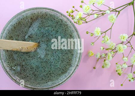 Körperpeeling mit Kaffee Draufsicht. Körperpeeling in einem Kunststoffbehälter auf einem rosa Hintergrund mit einem Zweig Gipsophila. Gel mit zerkleinerten Aprikosengruben. Stockfoto