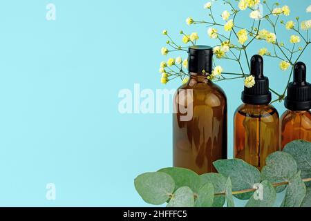 Kosmetikflaschen in verschiedenen Formen auf blauem Hintergrund mit Platz für Text. Drei braune Glasflaschen mit einem Zweig aus Gypsophila und Eukalyptus. se Stockfoto