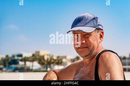Lächelnder Mann mittleren Alters, der eine Mütze trägt und an einem tropischen Strand sitzt Stockfoto