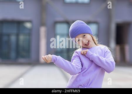 Nettes kleines kaukasisches Mädchen acht Jahre alt mit blonden Haaren lächelnd im Freien. Kind trägt stilvolles Hemd und Strickmütze in violetter Farbe. Trendige Farbe von Stockfoto