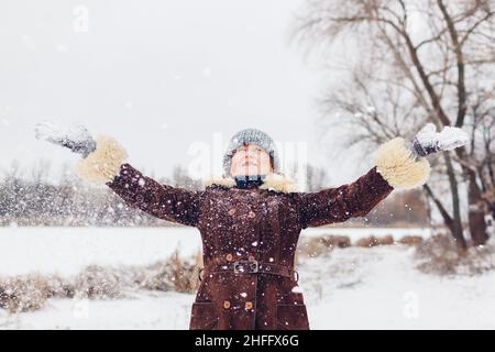 Porträt einer glücklichen älteren Frau, die Schnee in einen verschneiten Winterpark wirft. Spaß beim Spielen im Park während des Spaziergangs. Saisonale Aktivitäten Stockfoto