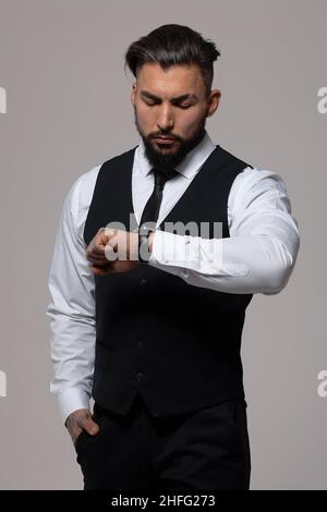 Bärtiger hispanischer Kerl in dunkler Weste und weißem Hemd mit Krawatte, der im Studio mit Händen in Taschen auf die Kamera blickt Stockfoto