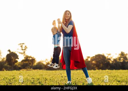 Mutter und ihr Kind spielen im Freien zusammen. Mutter und Sohn in Superhelden-Kostümen. Mama und Baby haben Spaß, lächeln und umarmen. Familienurlaub und Zweisamkeit Stockfoto