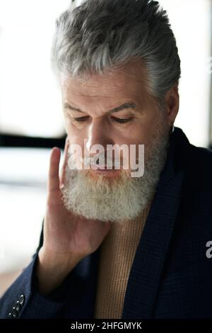 Nahaufnahme eines eleganten, nachdenklichen, grauhaarigen Mannes mittleren Alters, der nach unten blickt und seinen Bart berührt, während er in einem Gebäude posiert Stockfoto