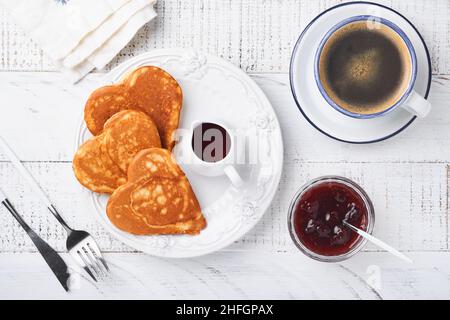 Pfannkuchen mit Beerenmarmelade und Honig in Herzform und heiße Tasse Kaffee auf weißem Holzhintergrund. Konzept Frühstück für Valentinstag oder pleasa Stockfoto