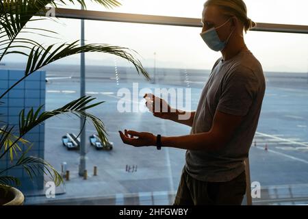 Mann, der am Flughafen oder an öffentlichen Orten Desinfektionsmittel auf die Hände sprüht. Hintergrundbeleuchtung vom Fenster, Silhouette. Mann in Gesichtsmaske, Flug und Covid Sicherheit Stockfoto