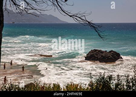 Glyfada, Korfu, Griechenland - 06. August 2021: Nicht erkannte Menschen schwimmen im Meer mit wunderschönen Wellen, die auf Felsen krachen und Wasser auf die Felsen spritzt Stockfoto