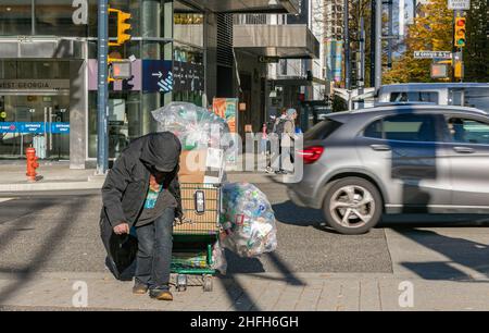 Obdachloser alter Mann auf einer Straße in Vancouver, BC, Kanada. Ein obdachloser Mensch kämpft einen Hügel hinauf, während er seinen Behälter mit Dosen trägt und seinen Trolley-N schiebt Stockfoto