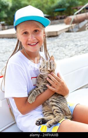 Weißes Teenager-Mädchen in Baseballmütze mit freundlichem Gesicht hält sanft tabby Kätzchen, Blick auf die Kamera, lächelnd, im Boot sitzen. Kleine niedliche Katze schnurrt ein Stockfoto