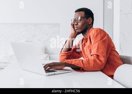 Der müde, zweifelende afroamerikanische Mann passt eine stilvolle Brille an, die am Tisch mit einem modernen Laptop in hellem Raum sitzt Stockfoto