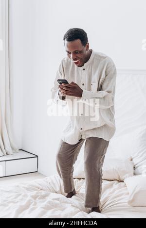 Ein lustiger, aufgeregter junger afroamerikanischer Mann in legerer Kleidung springt in einem modernen Zimmer mit Mobiltelefon auf einem großen Bett hoch Stockfoto