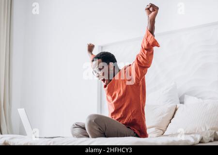 Fröhlicher afroamerikanischer Kerl in legerer Kleidung und Brille legt die Hände hoch, die in der Nähe eines offenen Laptops auf einem bequemen Bett zu Hause sitzen Stockfoto