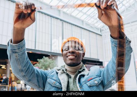Emotional glücklicher afroamerikanischer Männerfotograf in Denim-Jacke blickt in hell erleuchteter, extremer Nahaufnahme auf Vintage-Film Stockfoto