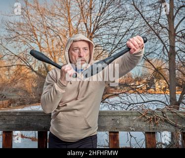 Outdoor-Porträt des älteren Mannes (Ende 60s) Training mit indischen Clubs in seinem Hinterhof, Winternachmittag, Fitness über 60 Konzept Stockfoto