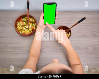 Frau, die Tabbouleh in einer Holzschüssel mit Essstäbchen isst und auf den grünen Bildschirm ihres Mobiltelefons schaut Stockfoto