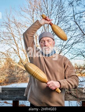 Älterer Mann (Ende 60s) trainiert mit hölzernen indischen Clubs in seinem Hinterhof, Winternachmittag, Fitness über 60 Konzept Stockfoto