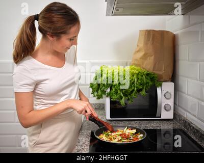 Junge Frau, die gerne zuschaut, wie Gemüse auf dem Cerankochfeld in der Küche gekocht wird, Stockfoto