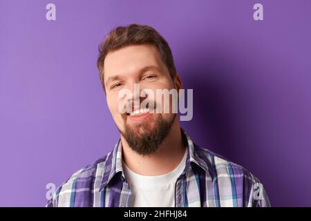 Glücklicher bärtiger Mann im Hemd lächelnd und schaute auf die Kamera auf purpurem Hintergrund. Junger, hübscher und stilvoller Kerl Stockfoto