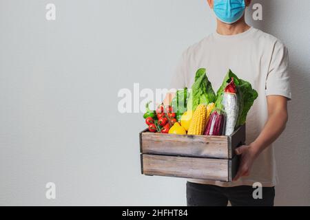 Junger Mann in schützender Gesichtsmaske, der Holzkiste in den Händen hält. Mann Kurier mit Shopping-Box. Sichere Online-Lieferung vom Supermarkt nach Hause Stockfoto