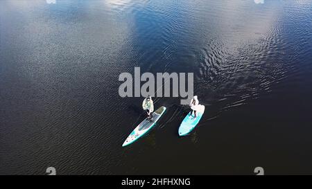 Draufsicht auf ein Paar, das auf dem See Paddle-Boarding macht, Sport- und Lifestyle-Konzept. Luftaufnahme eines Mannes und einer Frau, die auf einem Surfbrett stehen und mit einem rudern Stockfoto