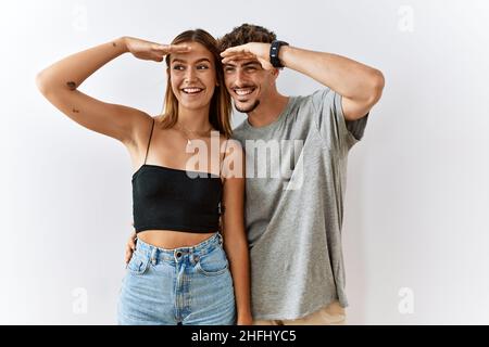 Junges schönes Paar, das über einem isolierten Hintergrund zusammensteht, sehr glücklich und lächelnd und weit weg mit der Hand über dem Kopf schaut. Suchkonzept. Stockfoto
