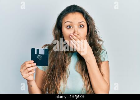 Junge hispanische Mädchen hält Diskette, die den Mund mit der Hand bedeckt, schockiert und Angst vor Fehlern. Überraschter Ausdruck Stockfoto